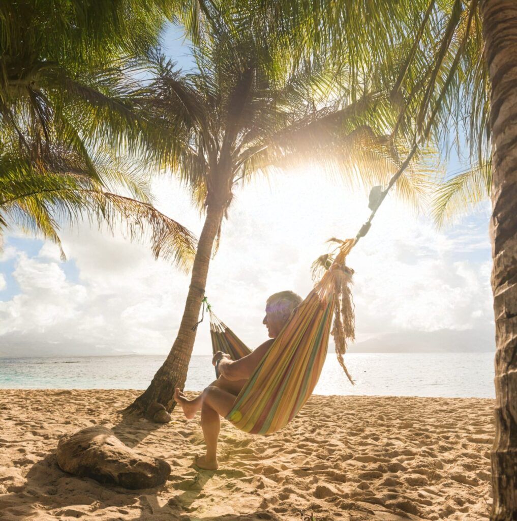 Retired man relaxing in a colorful hammock under palm trees on a sunny beach in Panama, with the ocean and clear sky in the background.